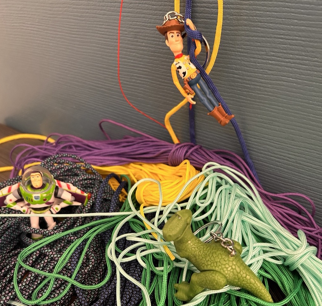 Figuras de un vaquero, un astronauta y un dinosaurio de juguete entre montañas de cordón paracord de colores